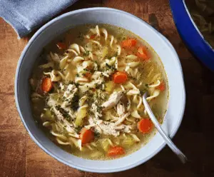 A steaming bowl of rotisserie chicken noodle soup topped with fresh herbs, showcasing tender chicken, colorful vegetables, and hearty noodles in a flavorful broth.